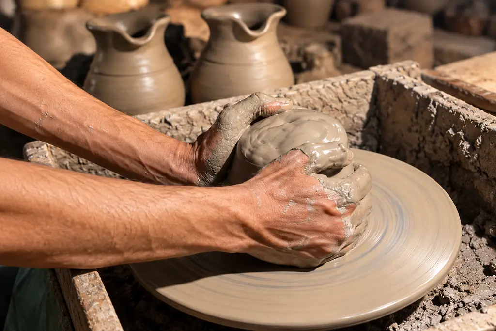 An artisan’s clay-covered hands centring a mound of wet clay on a potter’s wheel, freshly thrown jugs drying behind.